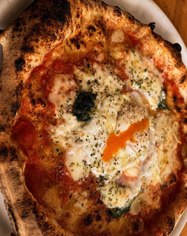 A bismarck Neapolitan pizza at Frey's Famous — a blistered, leopard-charred cornicione around molten mozzarella, basil, and a runny orange egg yolk broken across the center.