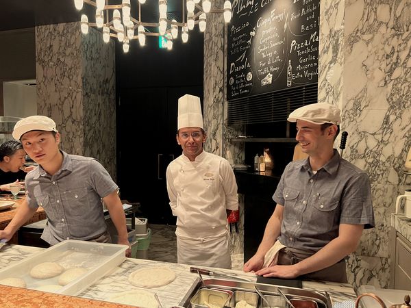 Three pizzaioli behind the marble counter of an open pizza kitchen, dough balls and topping trays before them, a handwritten chalkboard menu and a row of hanging lights behind.