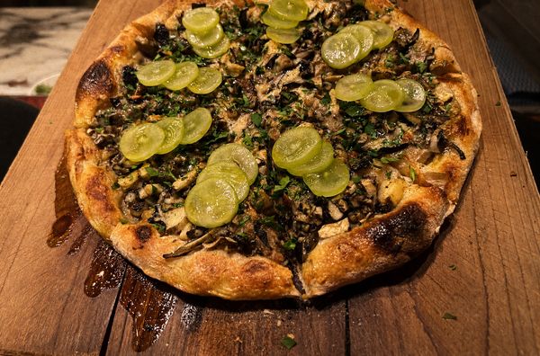 A wood-fired pizza on a serving board topped with mushrooms, chopped herbs, and translucent pale-green shine-muscat grape slices, a honey glaze pooling on the board.