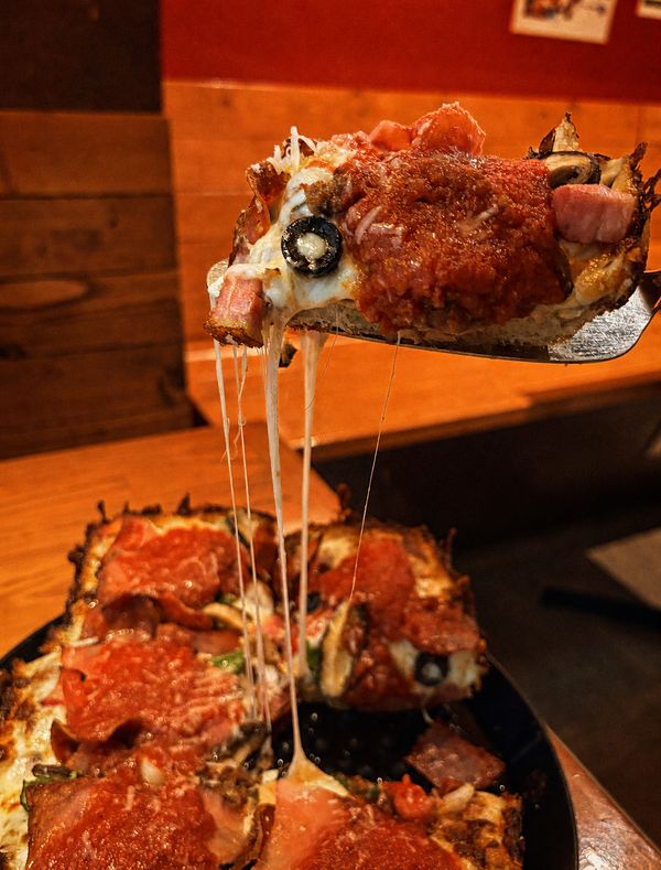 A square Detroit-style slice lifted from the pan at Pizzakaya, mozzarella stretching in long strands, topped with pepperoni, ham, black olives, and a stripe of chunky tomato sauce.
